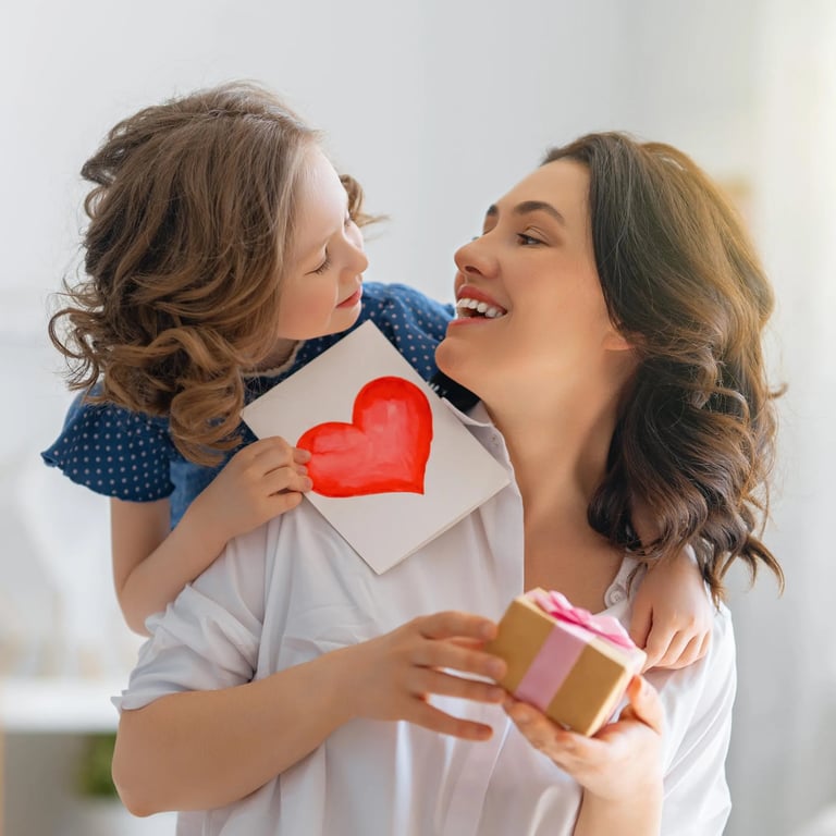 Kind umarmt Mama von hinten mit Bild in der Hand, auf dem ein aufgemaltes Herz ist. Mama hat bereits kleines Geschenk in der Hand.